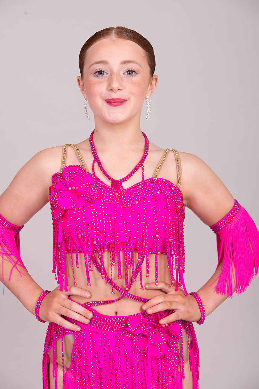 (BUDGET)11 Rose Pink Competition Latin Dance Dress. Flower detail to the chest & Hip. Fringed skirt with open side detail. Fringed arm cuffs & detailed necklace. Joined by crossover straps