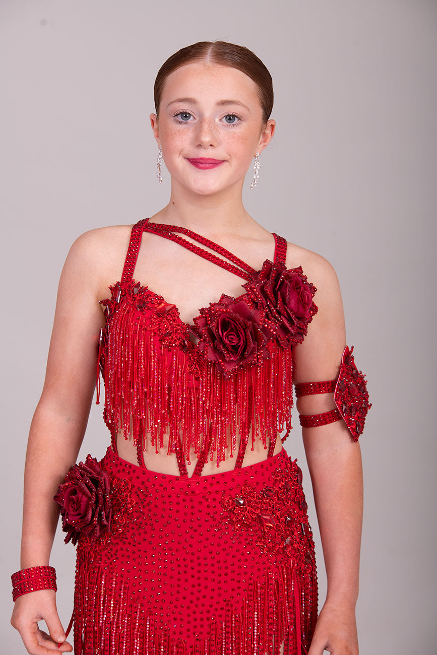 (BUDGET)13 Deep Red Competition Latin Dance Dress. Full of Red stoned fabric tassels & stoned motifs on the Bra style top. Heavily stoned flowers & strap detailing finish the top off. Skirt has flower to the Hip with stoned motifs and fabric strips over.